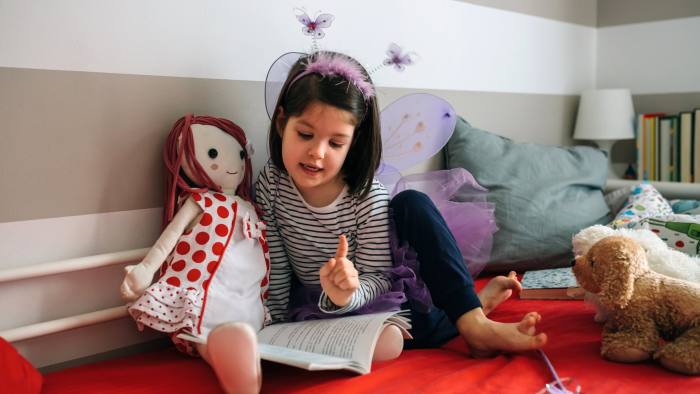 Little girl disguised as a butterfly sitting on the bed reading a book to her rag doll