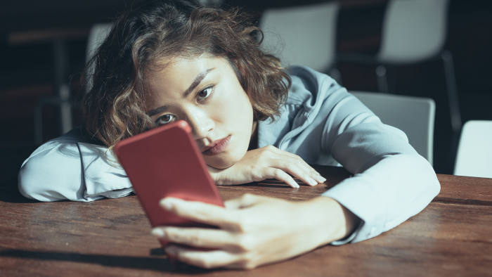 Closeup of thoughtful young Asian woman leaning on table and surfing Internet on red mobile phone at cafe. Female office worker browsing news feed during break. Mobile communication concept