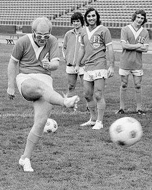 Elton John, co-owner of the LA Aztecs in the 1970s, takes a kick in front of star player George Best