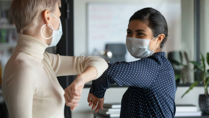 Smiling diverse female colleagues wearing protective face masks greeting bumping elbows at workplace, women coworkers in facial covers protect from COVID-19 coronavirus in office, healthcare concept