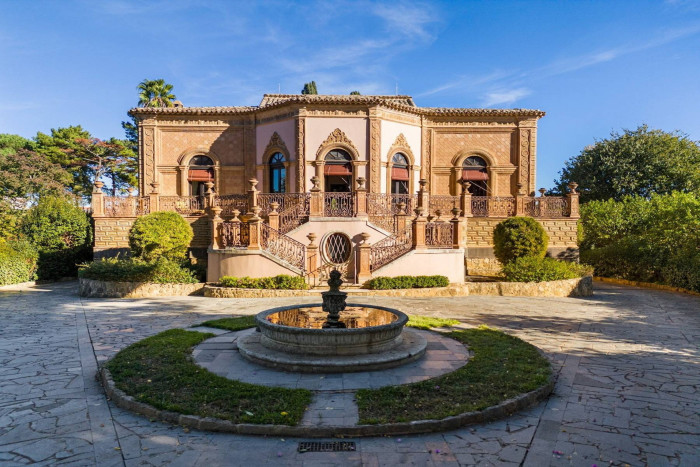 The highly decorate facade of a 19th-century Sicilian villa with a circular fountain in the driveway and steps lined with ornate railings leading to the entrance