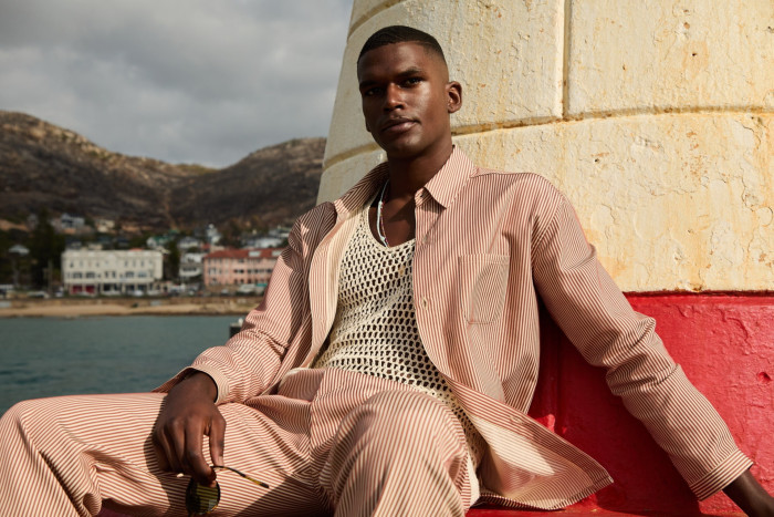 A model leans back on a sea wall. He wears a string vest and over it a peach and white striped shirt with matching trousers