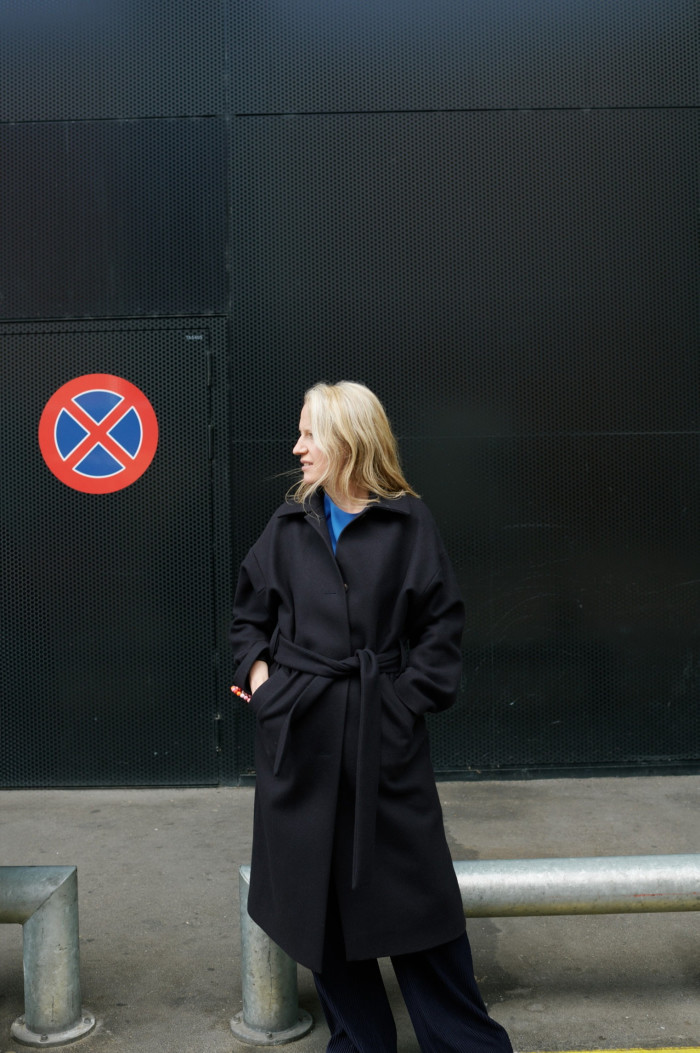 A middle-aged woman with long blond hair facing the side in a dark blue trench coat and a light blue jumper poses next to a ‘no stopping’ sign hanging on a black wall