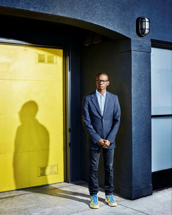 A man in a blue suit standing in front of a dark wall by a yellow door