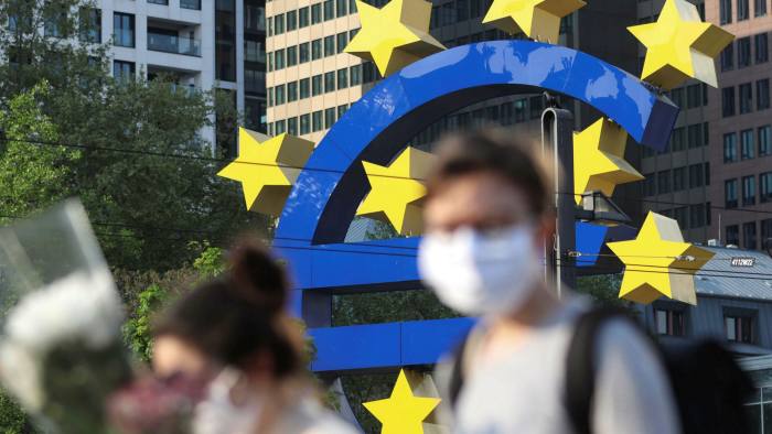 People wearing face masks walk in front of a big euro sign in Frankfurt, western Germany, as the European Central Bank headquarters can be seen in the background