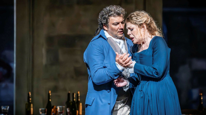 A man in a blue frock coat holds a woman in a blue dress as they both sing