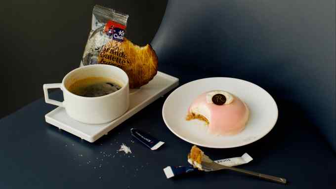 A plastic airline tray with a black coffee and a biscuit next to a rich an expensive-looking gateau on a china plate