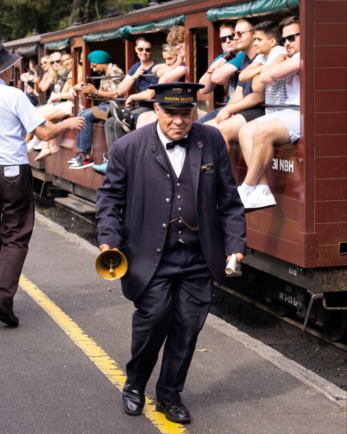 The station master at Belgrave station rings his bell on the platform by the Puffing Billy steam train, on which passengers are sitting with their legs hanging over the side
