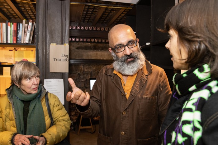 A middle-aged woman, a middle-aged man with a white beard and a young woman are captured talking in a gallery.