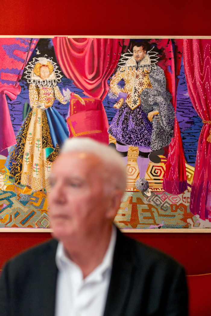 An old man dressed in a dark blazer and a white shirt is portrayed blurringly while sitting in front of a colourful painting of an aristocratic couple hanged in a red room.