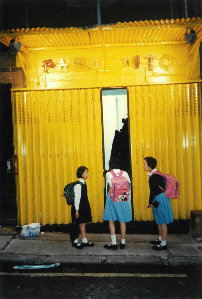 Three school girls with backpacks stand in front of a small building made of bright-yellow corrugated metal