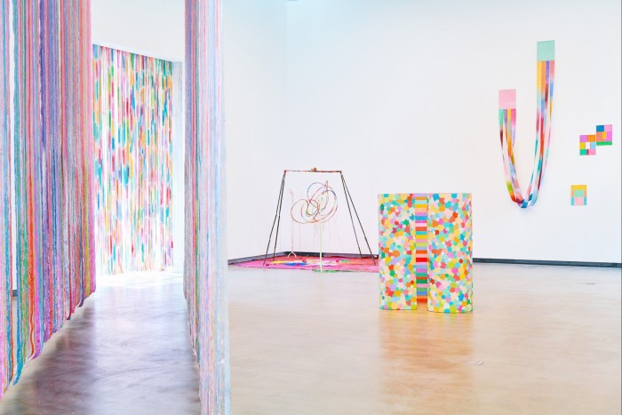 An installation view, with a tunnel-like structure made of long ribbons to the left of the image and smaller, colourful sculptures on the floor and wall to the right