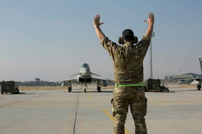 A man in military uniform with hands raised high, in front of a fighter jet