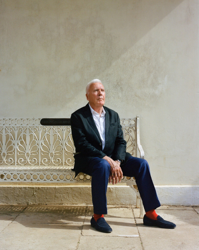 An old man dressed in a black blazer, white shirt and blue trousers and loafers stares into the distance to his left while sitting on a white iron bench decorated with floral motifs.