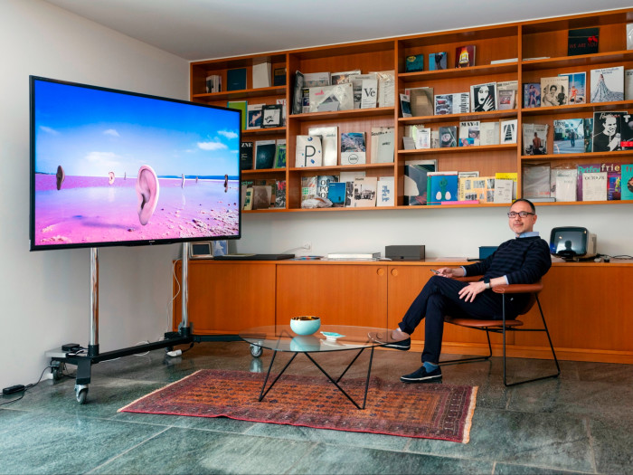 A middle-aged man dressed in a stripy dark jumper, blue trousers and glasses sits on a leather chair before a busy bookshelf. On the left of the scene, a wide TV screen shows a beach inhabited by floating ears