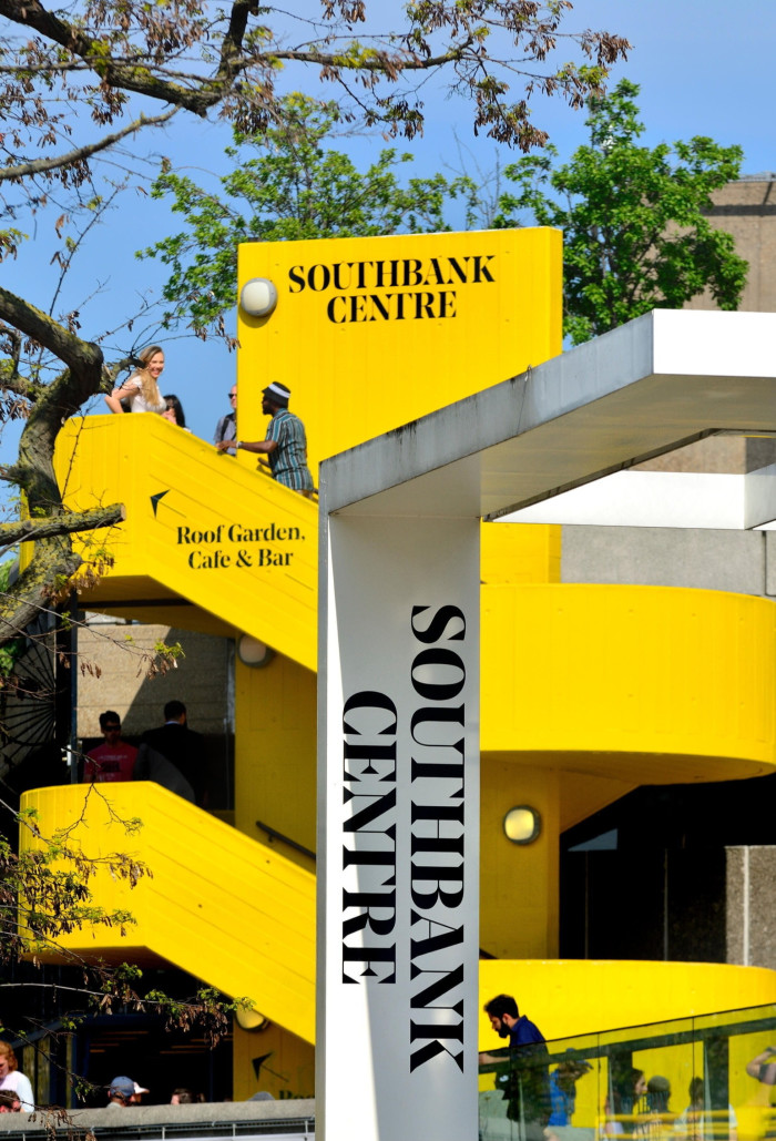 The vibrant yellow staircase of the Southbank Centre in London stands out against a clear blue sky. People are seen walking and talking on the steps. A large sign in the foreground reads “SOUTHBANK CENTRE”