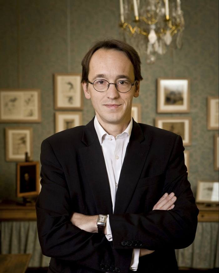 A man with short hair and glasses in a black jacket crossing his arms in a grand room with small pictures on the wall behind him