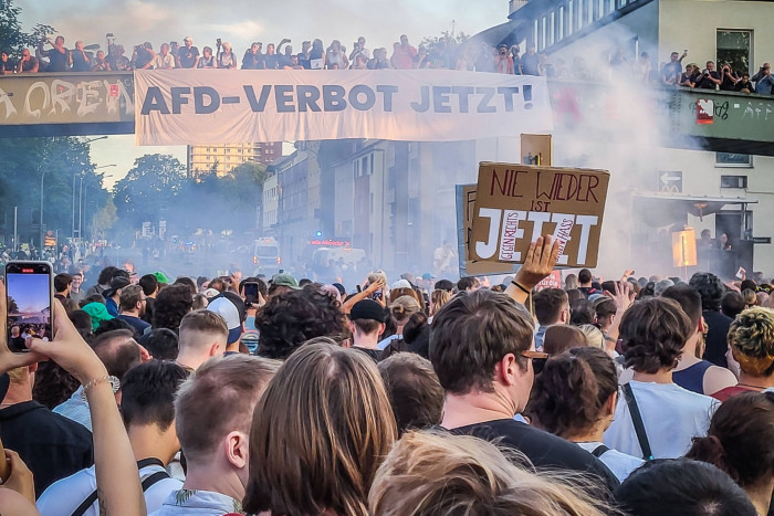 A large crowd in a smoky street. One person is holding up a mobile phone to take a photo, another holds a home-made banner with the words ‘Wieder ist Jetzt’, German for ‘Never again is now’