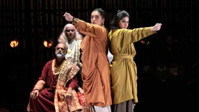 Two women stand back to back with their arms raised forward. One is wearing an orange robe, the other a golded on