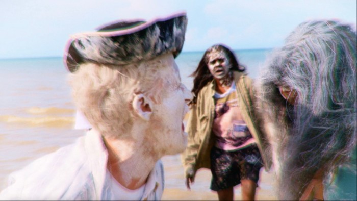 Three blurred figures covered in white face paint stand near the ocean