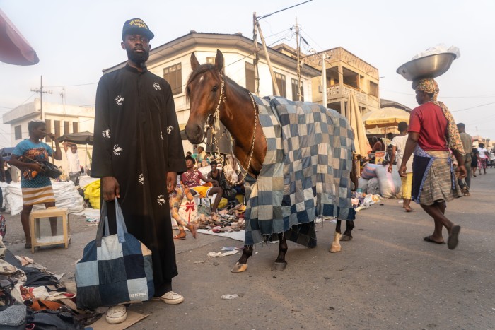 A man in a long robe and baseball hat stands next to a horse which wears a patchwork denim blanket