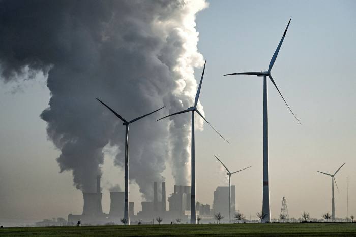 Wind turbines stand next to a power plant emitting large plumes of smoke