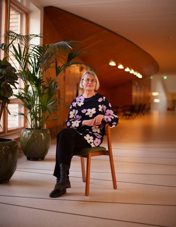 Full-body shot of Raija Kuokkanen, seated in what looks like a hallway and wearing a black floral top and black pants