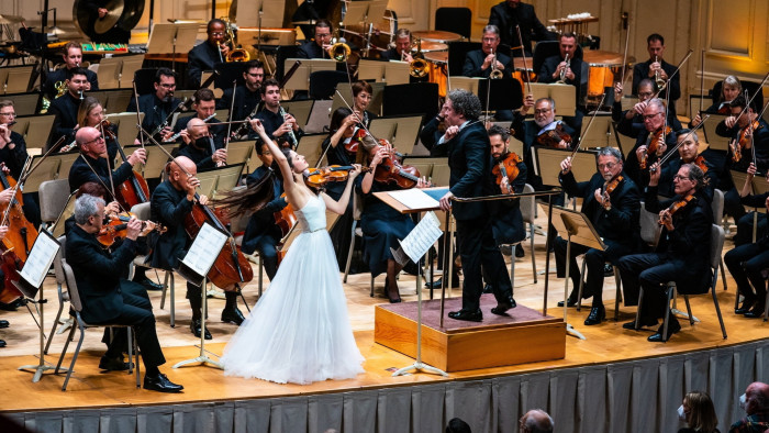 An orchestra performs on stage in front of an audience. All the musicians are dressed in black apart from a violin soloist, who is dressed in a white gown and performs at the front of the stage next to a male conductor.