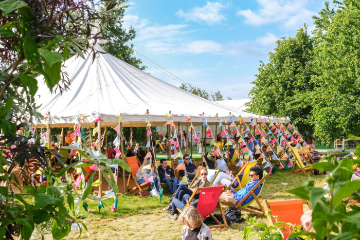 People sit in deckchairs in a field on a sunny day, near at tent whose ropes are decorated with bunting