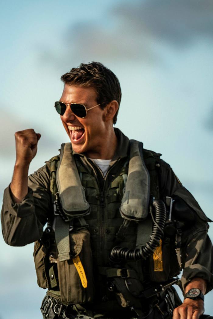 Tom Cruise in pilot suit and sunglasses, smiling, punches the air with one fist