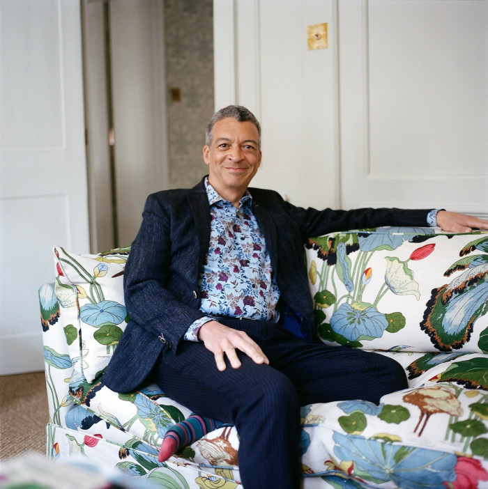 A man in a navy suit and floral shirt sits back smiling on a patterned sofa