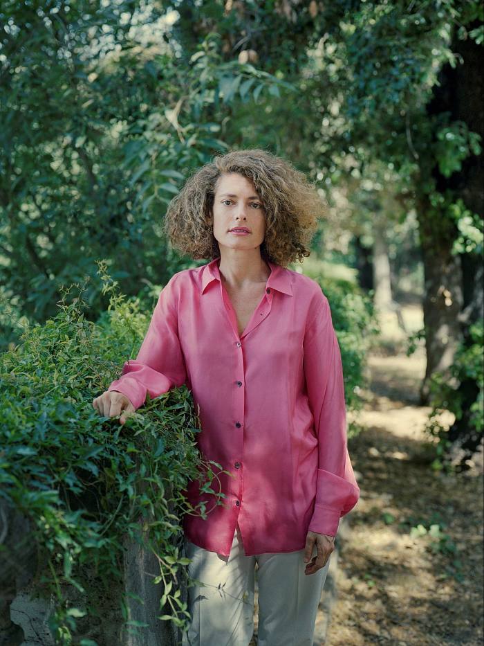 A woman with brown frizzy hair wearing a long pink shirt stands in the shade of a wood and rests her right arm on a bush