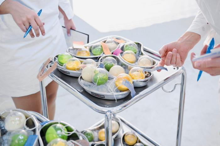 The hands of people in white lab coats pointing at metal dishes laden with transparent balls filled with thick white, green and yellow liquids