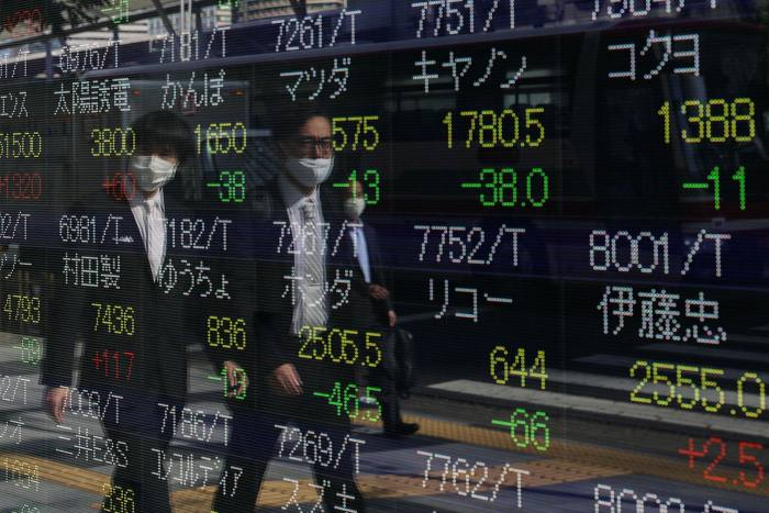 Pedestrians wearing face masks are reflected in an electronic screen showing stock market information on the street