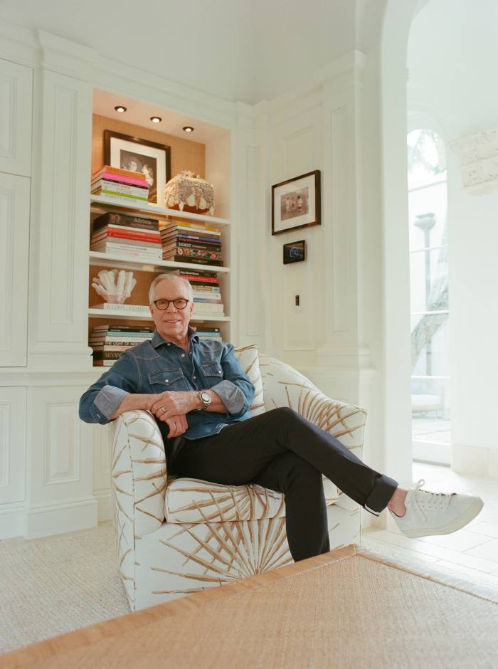 Hilfiger sits on an armchair in front of a bookshelf in his house