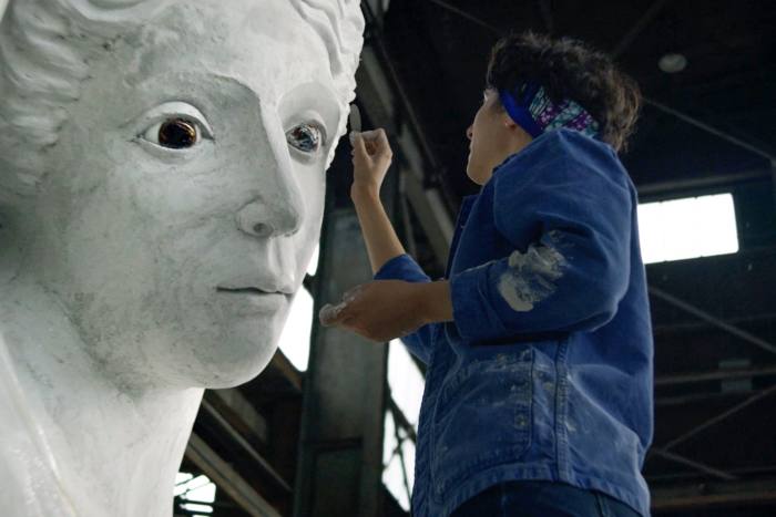 A woman in a blue cotton workman’s jacket sculpts a large woman’s face