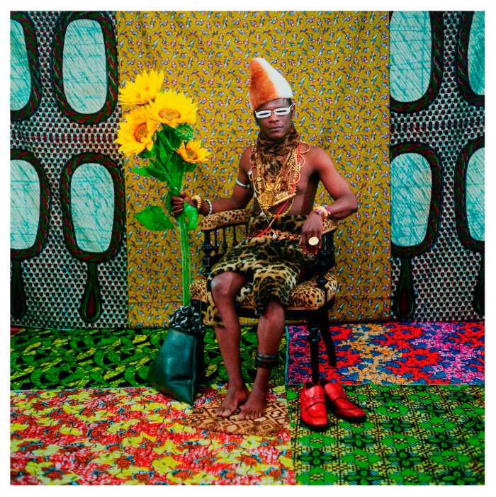 Photo of a man wearing leopard print sitting in a leopard print chair holding a bunch of sunflowers. On the floor and on the wall are bright fabrics