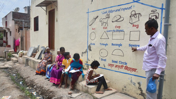 A schoolteacher in an Indian village points to pictograms and English words painted on the outside wall of a house while students are seated on the ground next to it with notebooks in hand