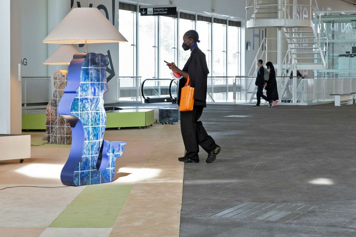 A woman in a suit walks across grey carpet towards a tall blue lamp sculpture