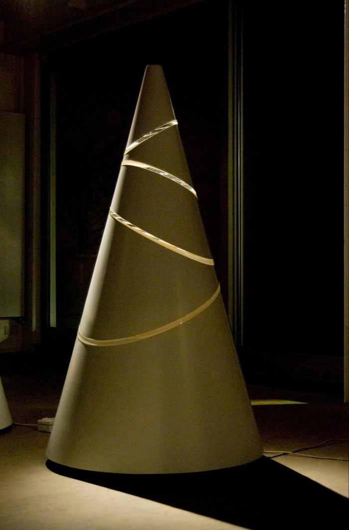 In a scarcely lit room, a yellow, cut-out cone stands tall in a bare setting, projecting a dark shadow on the floor