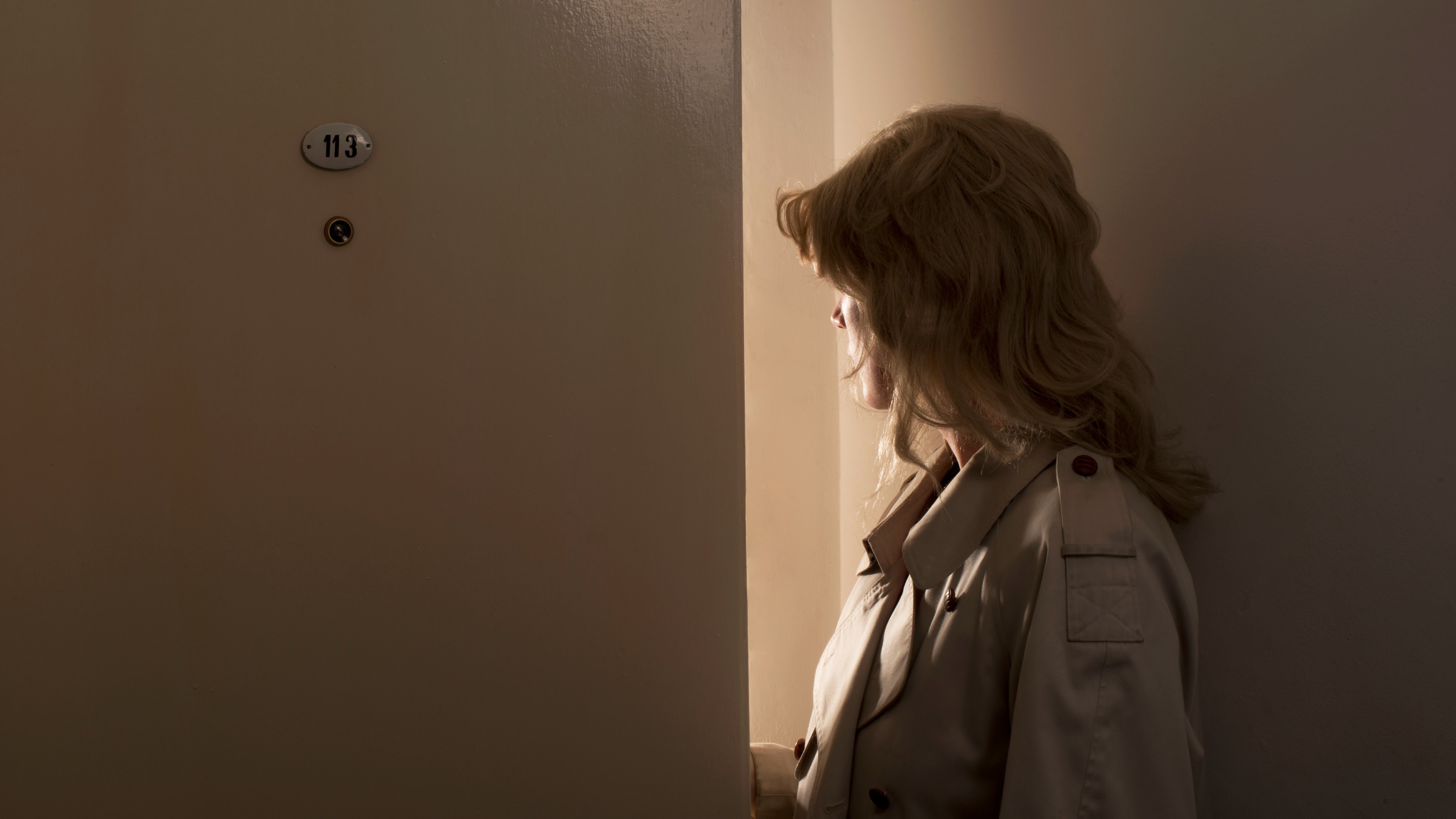 A woman in a trench coat looks behind a door