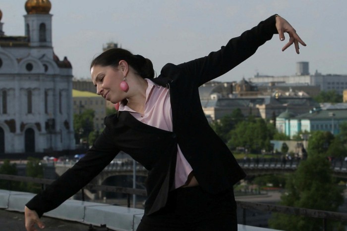 Sindeyeva, arms out wide, head to one side, is dancing. She is high up above the city, with the roofs of buildings behind her