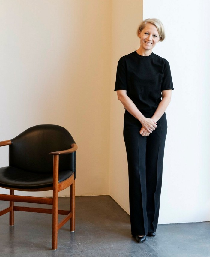 A woman in black stands, smiling, against a white wall next to a fancy chair