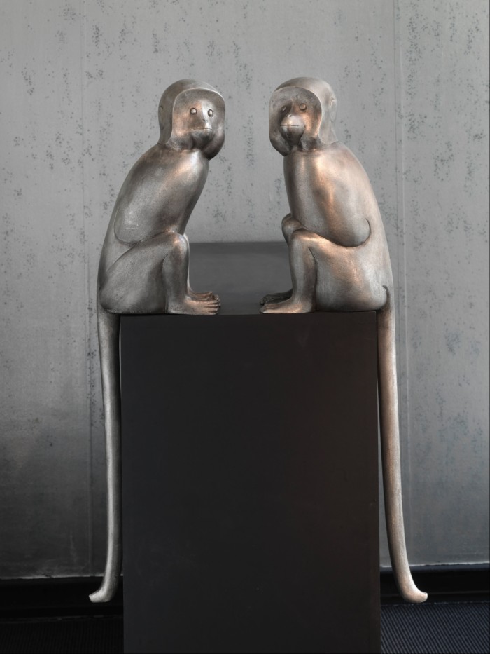 Two silver monkey sculptures facing each other on a black box, with long tails running to the floor