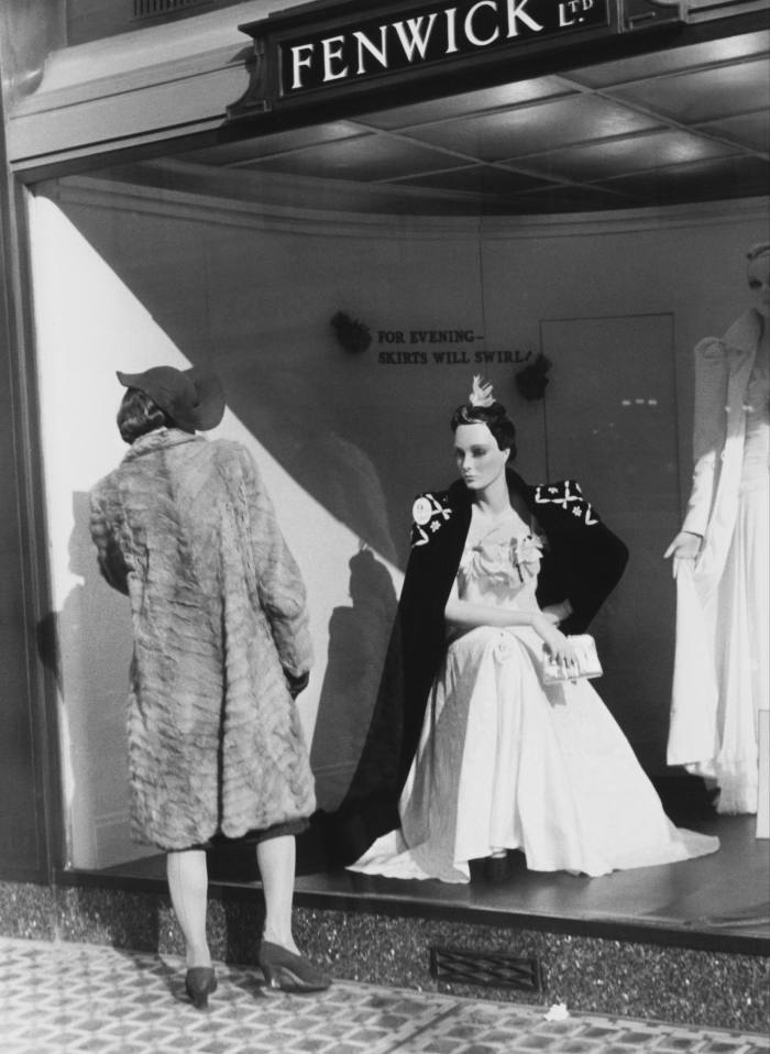 A woman in fur coat and hat looks at a mannequin dressed in an evening gown in a shop window