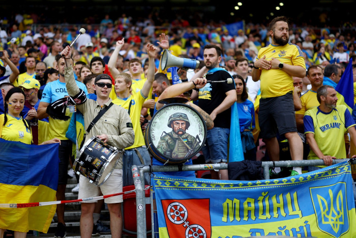 Two men with drums stand in front of a crowd of supporters, many of them dressed in yellow and blue. On the front of one of the drums is a painting of a soldier
