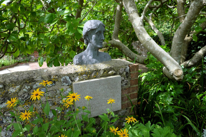 grave stone with inscription and sculpture of Virginia Woolf’s head, beneath a tree and surrounded by yellow flowers