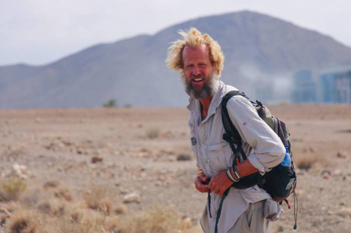 A man looking rough with scraggly beard in a blue shirt and backpack in front of some scrubland