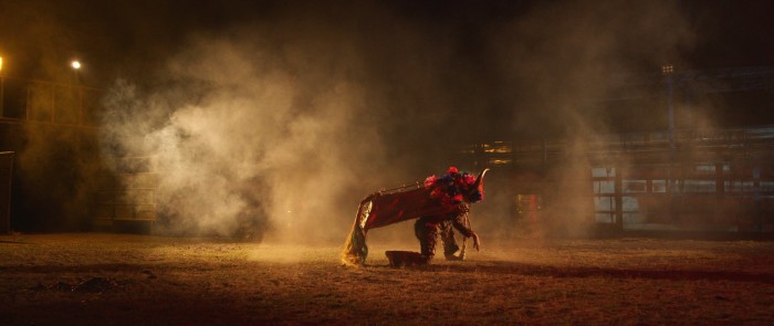 In a photograph, a person dressed in a bull costume with floral front decorations kneels in a dimly lit, dusty rodeo arena 