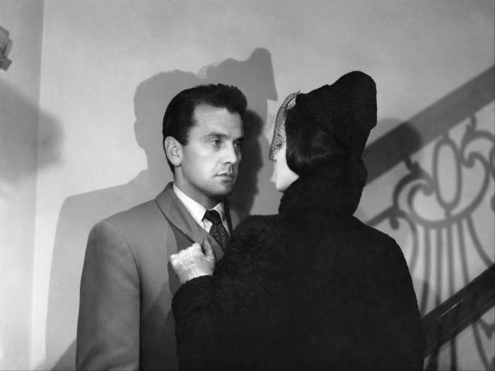 A woman in a black coat and hat played by Lucia Bose holds a man played by Massimo Girotti by the lapel on a staircase in ‘Story of a Love Affair’ 